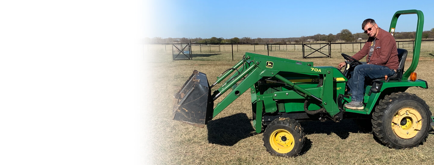 Male sitting upon green and yellow tractor with fence and grassy pasture in background.